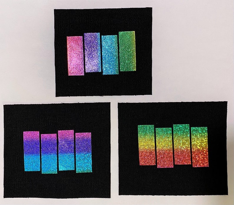 May include: Three black fabric patches with different colored iridescent stripes. The top patch has pink, purple, and blue stripes. The middle patch has pink, purple, and blue stripes. The bottom patch has green, yellow, and red stripes.