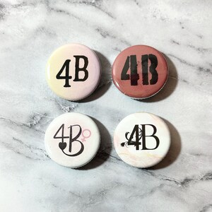 May include: Four round buttons with the letters "4B" in black on different colored backgrounds. The first button is yellow with a pink gradient, the second is brown, the third is white with a pink heart and female symbol, and the fourth is white with a pink marble pattern.