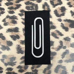 May include: A black rectangular patch with a white paperclip design. The patch is set against a background with a leopard print pattern in shades of brown and tan.