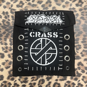 May include: A black fabric patch featuring white embroidered text that reads Dystopia at the top and Crass in the center above a circular logo. The patch includes six silver metal grommets along the sides and white stitching around the edges.