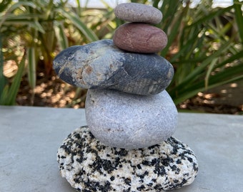 May include: A stack of five smooth, round stones in a zen garden setting. The stones are gray, black, and red, and are balanced on top of each other.