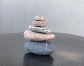 May include: A stack of five smooth, gray, brown, and pink stones balanced on top of each other on a black surface.