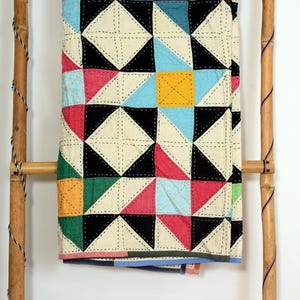 Könnte beinhalten: Ein rechteckiger Quilt mit einem geometrischen Muster aus Dreiecken in Schwarz, Creme, Gelb, Pink, Grün und Blau. Der Stoff ist durchgehend mit sichtbarem weißem Steppstich vernäht, was eine gitterartige Textur auf der bunten Oberfläche erzeugt.