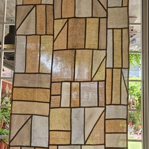 May include: A rectangular curtain with a patchwork design. The curtain features various rectangular and triangular shapes in shades of beige, tan, and brown, outlined with dark brown lines. The curtain hangs in front of a window, with a dark brown border at the top.