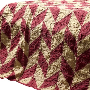 May include: A handmade quilt featuring a diamond pattern in burgundy and cream. The quilt is draped over a wooden surface, highlighting the texture and stitching. The colour scheme and design suggest a rustic or farmhouse style.