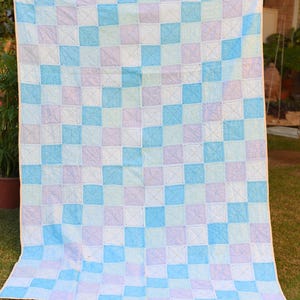May include: A handmade quilt with a checkered pattern in shades of blue, lavender, and white. The squares are stitched together with a contrasting thread, creating an X design. The quilt is displayed outdoors on a grassy area.