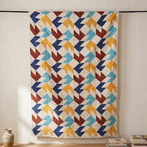 Op de afbeelding: Een rechthoekige quilt met een herhalend geometrisch patroon van pijlvormige vormen in marineblauw, geel, blauwgroen en kastanjebruin op een gebroken witte achtergrond. De quilt hangt aan een zwarte stang tegen een witte muur.