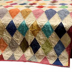 Op de afbeelding: Een handgemaakte quilt met een ruitpatroon in verschillende kleuren, waaronder rood, blauw, groen en oranje. De quilt is over een houten oppervlak gedrapeerd, wat de textuur en het ontwerp benadrukt. De afmetingen van de quilt zijn niet zichtbaar.