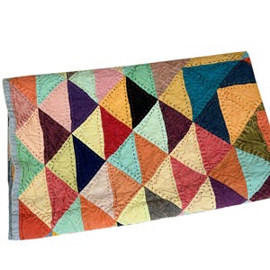May include: A colorful quilt with a geometric pattern of triangles in various shades of red, orange, yellow, blue, and purple. The quilt is folded and has a light blue border.
