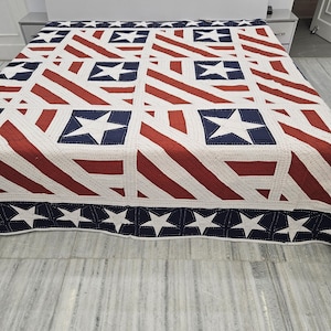 May include: A red, white, and blue quilt with a patriotic design. The quilt features alternating red stripes and white squares with blue squares containing white stars. The border is navy blue with white stars.