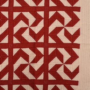 Könnte beinhalten: Eine textile Steppdecke mit einem sich wiederholenden geometrischen Muster aus dunkelroten und cremeweißen Formen. Der Stoff zeigt durchgehend sichtbare Steppnähte, die einen strukturierten Gittereffekt auf der gesamten Oberfläche erzeugen.