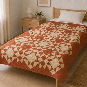 Könnte beinhalten: Eine Steppdecke mit geometrischem Muster in Rostorange und Cremeweiß. Das Textil zeigt ein sich wiederholendes Blockdesign mit Dreiecken und Quadraten, das über einem Bettrahmen liegt. Der Stoff weist eine deutlich sichtbare Steppstruktur auf.