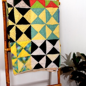 Könnte beinhalten: Eine rechteckige Patchwork-Decke mit einem Gitter aus dreieckigen Blöcken in verschiedenen Farben, darunter Schwarz, Weiß, Gelb, Türkis, Grün und Rot. Die Decke weist auf der gesamten Stoffoberfläche sichtbare, von Hand genähte Fäden auf.
