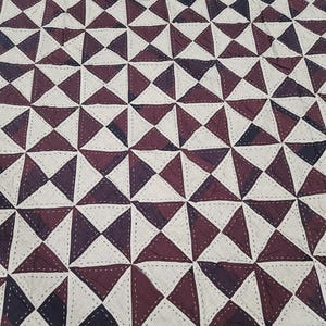 Könnte beinhalten: Nahaufnahme eines Textils mit geometrischem Muster. Das Design zeigt abwechselnd weiße und burgunderfarbene Dreiecke, wodurch sich ein sich wiederholendes Muster ergibt. Die Dreiecke sind mit weißem Faden zusammengenäht, was dem Stoff Textur und Details verleiht.