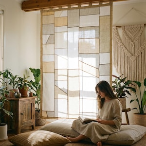 May include: A woman reading in a room with a large patchwork curtain. The curtain is made of beige, white, and tan fabric. The room is decorated with plants, a wooden cabinet, and a macrame wall hanging. The woman is wearing a light-coloured dress.