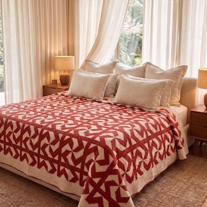 May include: A bed covered with a quilt featuring a repeating geometric pattern in deep red and off-white. The quilt has a stitched texture and hangs over the sides of the bed. Several off-white rectangular pillows are arranged at the head of the bed.