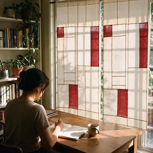 May include: A pair of floor-length curtains featuring a grid pattern of thin tan lines on a cream-colored fabric, accented with rectangular blocks of deep red. The curtains hang over a window, allowing soft light to filter through the weave.