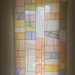 Puede incluir: Una cortina transparente con un diseño de patchwork geométrico. La cortina presenta formas rectangulares e irregulares en varios colores, incluyendo amarillo, azul, rosa y verde, delineadas con líneas marrones. La cortina cuelga frente a una ventana.