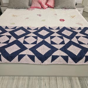 Peut inclure: Un lit avec une courtepointe blanche ornée de motifs floraux et d'un motif géométrique bleu et rose au pied. Des oreillers gris et roses sont disposés sur le lit. Le cadre de lit est gris.