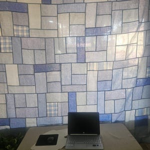 Pode incluir: Um laptop com tela preta está sobre uma superfície bege, ao lado de um mousepad e mouse pretos. O fundo apresenta uma cortina com um design de patchwork em tons de azul e branco.