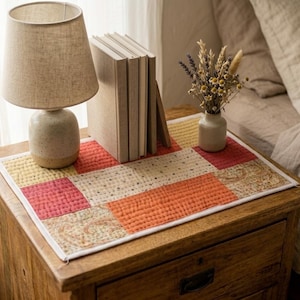 May include: A rectangular quilted fabric mat featuring a patchwork design of solid red, orange, and yellow squares alongside sections with a tan and brown paisley pattern, all bordered by a thin white trim and finished with visible stitching.