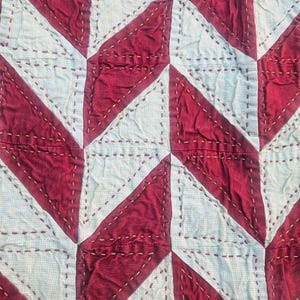Könnte beinhalten: Ein Textil mit einem sich wiederholenden geometrischen Muster aus abwechselnden dunkelroten und hellblaugrauen Dreiecken. Der Stoff ist mit sichtbaren, horizontalen Vorstichen in hellem Garn verbunden, was eine texturierte, gesteppte Optik erzeugt.