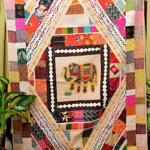May include: A vibrant, patchwork quilt featuring an embroidered elephant in the center. The quilt showcases a variety of colorful fabrics in a geometric design, with a cream-colored border and decorative stitching. The overall aesthetic is bohemian and handcrafted.