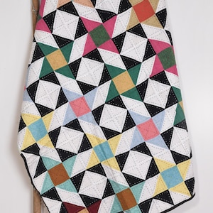 Puede incluir: Un edredón hecho a mano con un patrón geométrico. El edredón presenta cuadrados negros, blancos y coloridos en tonos de rosa, verde, amarillo y azul. El edredón está drapeado sobre una vara de bambú.