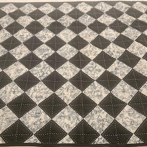 Könnte beinhalten: Eine gesteppte Decke mit einem sich wiederholenden Rautenmuster. Abwechselnde Rauten sind schwarz und weiß, wobei die weißen Rauten ein florales Design aufweisen. Die Decke ist mit einer schwarzen Bordüre eingefasst und mit weißem Faden genäht.