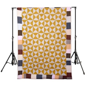 Könnte beinhalten: Eine handgefertigte Steppdecke mit einem geometrischen Muster aus gelben und weißen Dreiecken. Die Steppdecke hat einen Rand aus verschiedenfarbigen Quadraten, darunter Braun, Blau und Rosa. Die Steppdecke wird auf einem schwarzen Ständer vor weißem Hintergrund präsentiert.