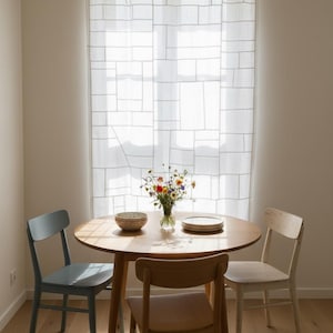 Pode incluir: Uma pequena área de jantar com uma mesa redonda de madeira, três cadeiras e uma cortina transparente. A mesa está posta com uma tigela, pratos e um vaso de flores. Uma cadeira é azul clara, as outras são de madeira.