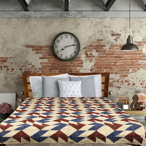 May include: A bed with a wooden headboard is covered with a quilt featuring a geometric pattern in cream, navy blue, and burgundy. The room has a brick wall, a large clock, and a black lamp.