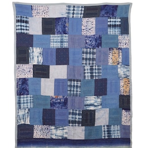 May include: A rectangular quilt featuring a patchwork of various blue fabric squares with diverse patterns including tie-dye, stripes, and solid shades. The quilt is finished with visible white running stitch embroidery and a solid grey border.