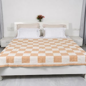 May include: A bed featuring a large quilt with a tan and white checkered pattern. The bed is dressed with two white rectangular pillows and two larger beige pillows. The bed frame is white and sits between two white bedside tables.