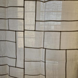 Puede incluir: Cortina blanquecina con un patrón geométrico de líneas grises oscuras. El diseño crea una apariencia de rejilla, con formas rectangulares y cuadradas. La tela es ligeramente translúcida, permitiendo el paso de la luz.