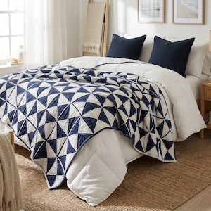 May include: A navy blue and white geometric patterned quilt covers a bed with two navy blue pillows. The quilt features a repeating triangle design. The bed is in a well-lit room with a neutral color palette.