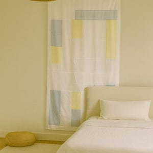 May include: A bedroom scene featuring a white bed with a matching pillow and duvet. A patterned curtain with blue and yellow rectangles hangs behind the bed. A woven pendant light and a round woven floor cushion complete the decor.