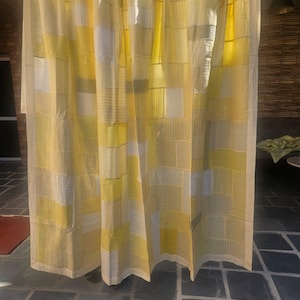 Puede incluir: Una cortina de retazos amarilla y blanca colgada de una barra. La cortina está hecha de varias piezas de tela rectangulares en tonos de amarillo, blanco y gris. La cortina cuelga frente a una pared de ladrillo y un suelo embaldosado oscuro.