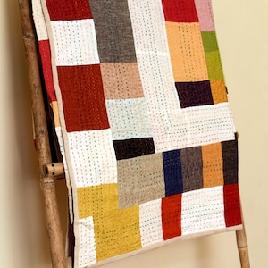 Może przedstawiać: Kolorowa patchworkowa kołdra z prostokątnymi kształtami w kolorach czerwonym, białym, żółtym, brązowym i niebieskim. Kołdra jest zawieszona na bambusowej drabinie, ukazując ręczne wykonanie i szczegóły szycia. Projekt łączy gładkie i wzorzyste tkaniny.