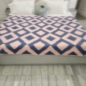 Può includere: Un letto con cuscini bianchi e una trapunta a motivi geometrici blu e rosa. La trapunta presenta un motivo a rombi ripetuto. Un piccolo cuscino decorativo con un fiore è anche sul letto. La struttura del letto è grigia.