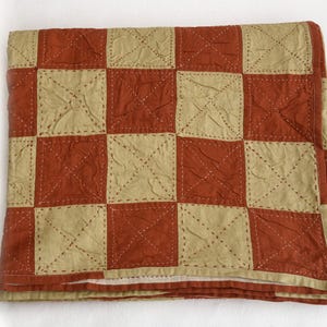 Op de afbeelding: Een opgevouwen quilt met een dambordpatroon. Afwisselende vierkanten zijn een gedempt rood-oranje en een lichtbeige. Elke vierkant heeft een gestikt X-ontwerp. De quilt is gemaakt van een textuurstof.