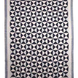 Könnte beinhalten: Eine rechteckige Steppdecke mit einem sich wiederholenden geometrischen Muster aus dunkel marineblauen und weißen Dreiecken. Das zentrale Design ist von einem dünnen, hellblauen Rand mit einem dezenten Kreuzstichmuster an den Außenkanten umrahmt.