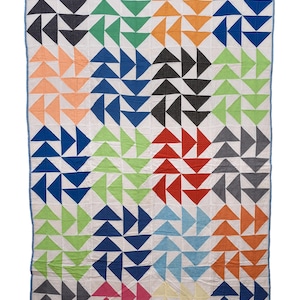 Puede incluir: Una colcha rectangular con un patrón repetitivo de triángulos multicolores dispuestos en bloques sobre un fondo blanco. La colcha está bordeada por un ribete de tela azul claro y muestra una variedad de colores que incluyen rojo, azul, verde, naranja, rosa, amarillo y gris.