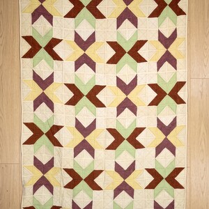 Könnte beinhalten: Ein rechteckiger Quilt mit einem sich wiederholenden geometrischen Sternmuster in den Farben Dunkelrot, Hellgrün, Gelb und Violett auf cremefarbenem Grund. Die Stoffoberfläche weist durchgehend sichtbare, gitterartige Steppnähte auf.