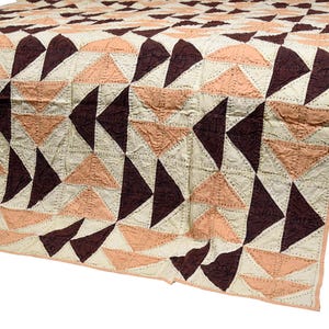 Pode incluir: Um edredom com padrão geométrico em creme, pêssego e castanho escuro. O edredom apresenta um design de triângulo repetido, com cada triângulo costurado. O edredom é drapeado sobre uma superfície de madeira escura.