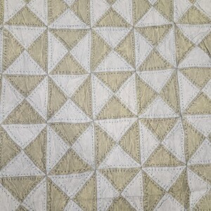 Könnte beinhalten: Nahaufnahme eines gesteppten Stoffes mit geometrischem Muster. Das Design zeigt abwechselnd Dreiecke in Off-White und Beige, die mit einer gepunkteten Linie zusammengenäht sind. Die Gesamtstruktur wirkt weich und leicht zerknittert.
