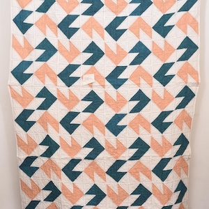 Könnte beinhalten: Eine handgefertigte Steppdecke mit einem geometrischen Muster. Das Design zeigt teal-, pfirsich- und weiße Formen, die in einem sich wiederholenden Muster angeordnet sind. Die Steppdecke wird auf einem Holzständer ausgestellt.