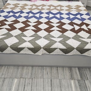 May include: A bed covered with a geometric patterned quilt in white, brown, blue, and green. The quilt is adorned with diamond and zig-zag shapes. Gray and pink pillows are arranged on the bed.