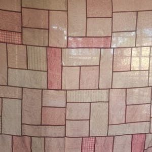 Puede incluir: Un textil de patchwork en tonos rosa, beige y crema, con líneas marrón oscuro que definen formas rectangulares y cuadradas. Algunos cuadrados presentan un patrón de cuadros. La tela parece ser lino o una fibra natural similar.