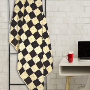 May include: A black and cream-coloured quilt with a diamond pattern hangs on a black metal ladder. The quilt is made of stitched squares. The background is a white brick wall. A red mug and laptop are on a white desk.
